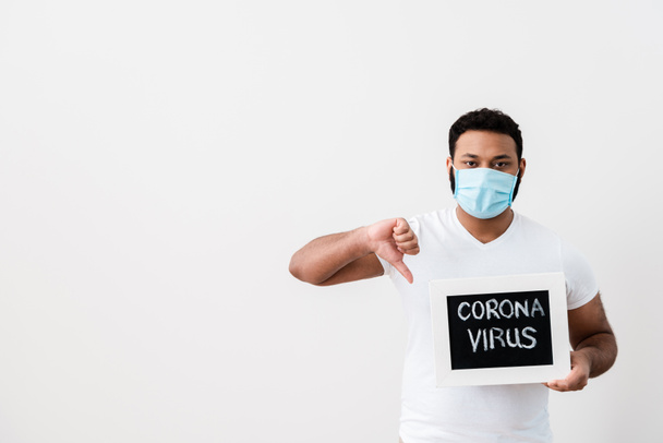 africký Američan v lékařské masce drží tabuli s koronavirem nápisy a ukazující palec dolů u bílé zdi  - Fotografie, Obrázek