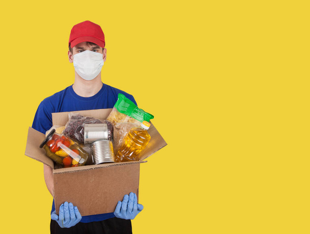 Young man, a volunteer in a medical mask and rubber gloves holds a cardboard box with humanitarian aid products on a yellow background. Social assistance. donation - Photo, image