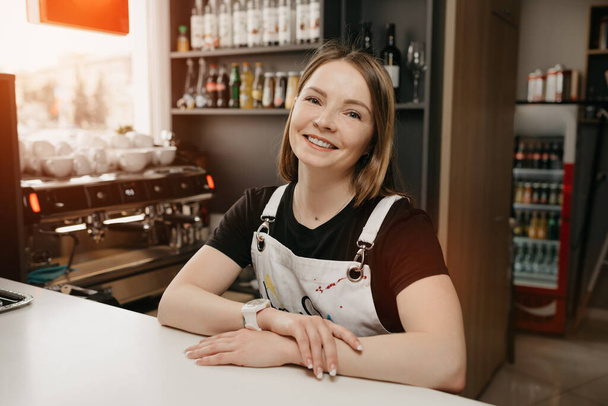 Kobiecy barista w białym fartuchu opiera się o barek w kawiarni. Ładna właścicielka firmy uśmiechnięta i stojąca ze złożonymi rękami w kawiarni. - Zdjęcie, obraz