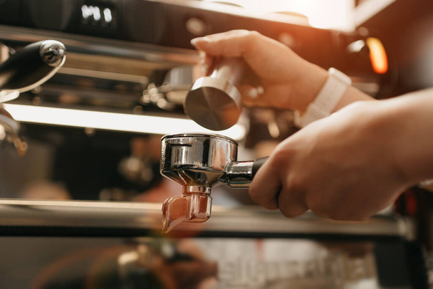 Een close foto van vrouwelijke handen met een metalen sabotage en een portafilter met koffie in een koffieshop. Een barista die zich voorbereidt op het persen van gemalen koffie voor het zetten van espresso of americano in een café. - Foto, afbeelding