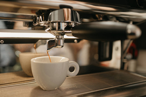 Ο Εσπρέσο ζυθοποιείται σε μια επαγγελματική μηχανή εσπρέσο σε μια καφετέρια. Ένα κοντινό πλάνο ενός καφέ που χύνεται σε ένα λευκό φλιτζάνι από μια καφετιέρα σε ένα καφέ. - Φωτογραφία, εικόνα