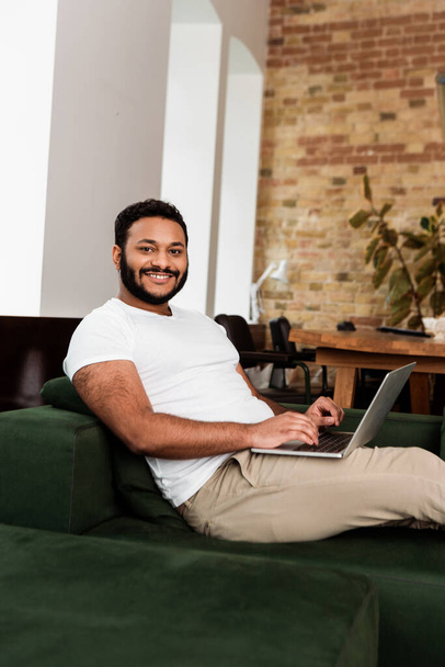 wesoły afrykański amerykański freelancer siedzi na kanapie z laptopem i patrzy w kamerę  - Zdjęcie, obraz