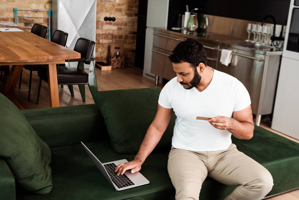bearded african american man holding credit card near laptop while online shopping in living room  - Photo, Image