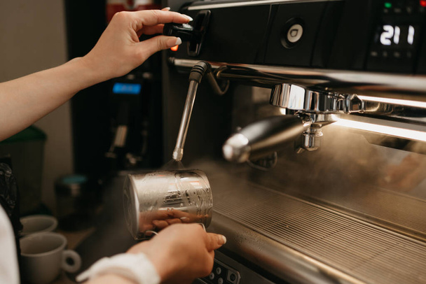 A female barista wears a white apron purges a metal mug with steam in a professional espresso machine in a coffee shop. A close-up photo of cleaning a cup in a coffee machine in a cafe. - Foto, Imagen