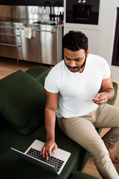 Afrikanisch-amerikanischer Mann hält beim Online-Shopping im Wohnzimmer Kreditkarte in der Nähe von Laptop  - Foto, Bild