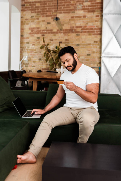 Afrikanisch-amerikanischer Mann hält beim Online-Shopping im Wohnzimmer Kreditkarte in der Nähe von Laptop mit leerem Bildschirm  - Foto, Bild