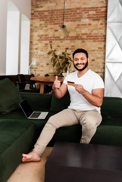 cheerful african american man holding credit card near laptop with blank screen while showing thumb up  - Photo, Image