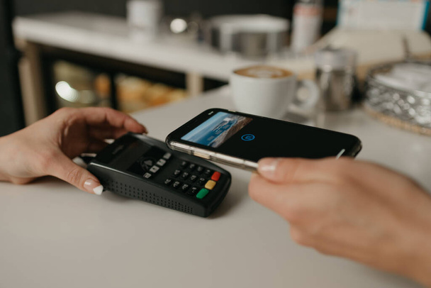 Eine Frau, die ihren Kaffee in einem Café mit einem Smartphone per kontaktloser PayPASS-Technologie bezahlt. Eine Barista hält einem Kunden ein Terminal zur Bezahlung hin. - Foto, Bild