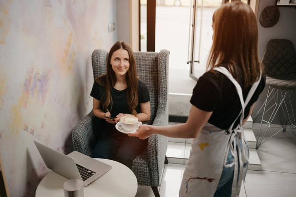 Uma empregada de bar oferece um café com leite a uma rapariga num café. Uma mulher com cabelos longos trabalhando remotamente em um laptop mantém distância social agarra uma xícara de café em um café
.  - Foto, Imagem