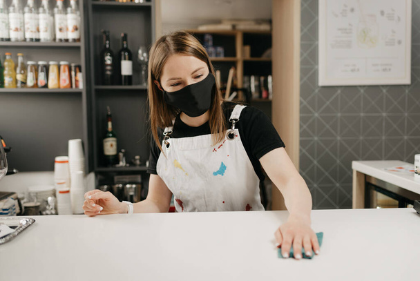 Een vrouwelijke barista met een medisch zwart gezichtsmasker wrijft in een koffieshop over de bar. Een mooie serveerster desinfecteert een café.. - Foto, afbeelding