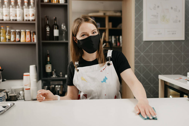 Kobieta barista w medycznej czarnej masce uśmiecha się i sprząta bar w kawiarni. Ładna kelnerka dezynfekująca kawiarnię.. - Zdjęcie, obraz
