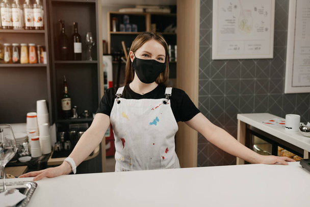 Μια χαρούμενη μπαρίστα με μαύρη μάσκα χαμογελά και περιμένει πελάτες στην καφετέρια. Μια όμορφη γυναίκα ιδιοκτήτρια του καφέ ποζάρει με τα χέρια της ορθάνοιχτα πίσω από το μπαρ.. - Φωτογραφία, εικόνα