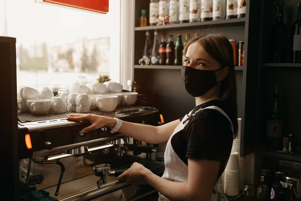 A happy female barista in a medical black face mask makes a cup of coffee in a cafe. A waitress posing near a coffee machine.  - Fotó, kép