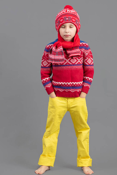 A child in warm clothes. Boy in a winter hat, scarf and red sweater barefoot. Fashionable boy in the studio. - Foto, afbeelding