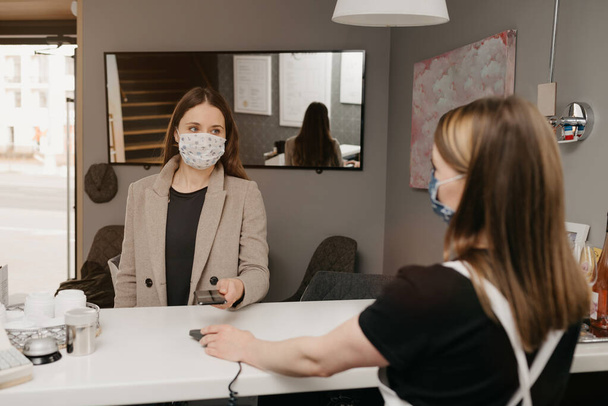 Ένα κορίτσι που φοράει μάσκα ιατρικού προσώπου χρησιμοποιεί ένα κινητό για να πληρώσει με τεχνολογία ανέπαφων πληρωμών. Μια γυναίκα μπαρίστα με μάσκα προσώπου κρατά σε πελάτη ένα τερματικό NFC για την πληρωμή. - Φωτογραφία, εικόνα