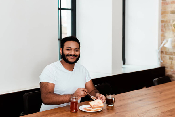 vidám afro-amerikai férfi kezében kanál mellett üveg édes lekvár és pirítós kenyér tányéron - Fotó, kép
