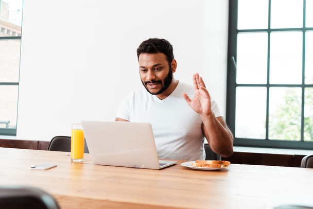 selective focus of bearded african american man waving hand while having video call near tasty breakfast on table - Photo, Image