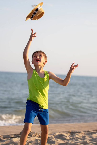 Boldog gyermek a tengeren. A fiú a parton feldobta a kalapját.. - Fotó, kép