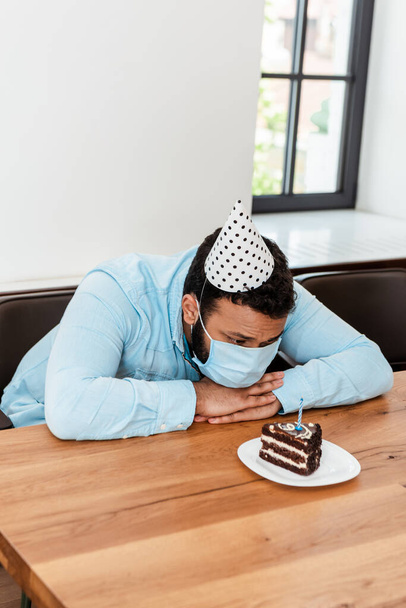 Afrikanisch-amerikanischer Mann in Partymütze und medizinischer Maske feiert Geburtstag allein und schaut auf Geburtstagstorte - Foto, Bild