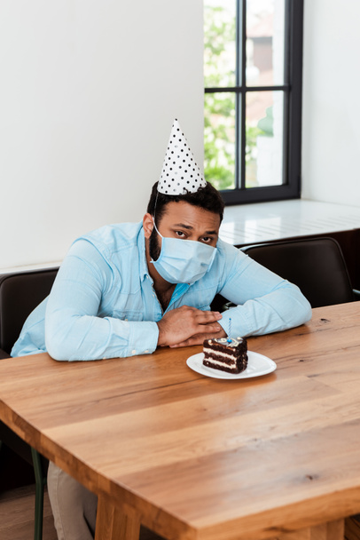 Afrikaan amerikaanse man in party cap en medisch masker vieren verjaardag alleen in de buurt van smakelijke verjaardagstaart - Foto, afbeelding