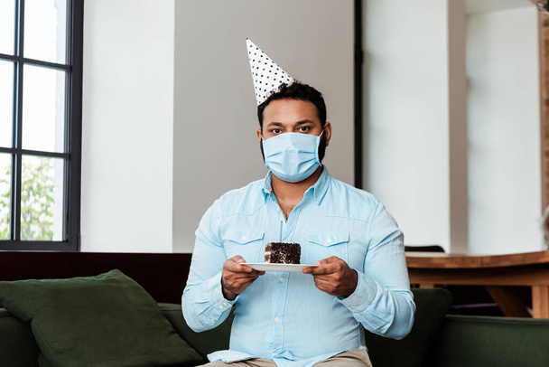 african american man in party cap and medical mask holding plate with birthday cake - Fotografie, Obrázek