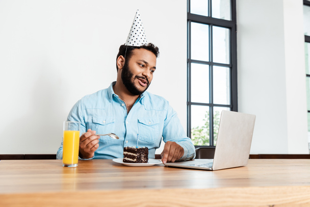 african american man in party cap looking at laptop while having video call near tasty birthday cake and glass of orange juice - Photo, Image