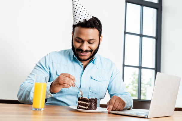 cheerful african american man in party cap looking at tasty birthday cake near laptop and glass of orange juice - Photo, Image