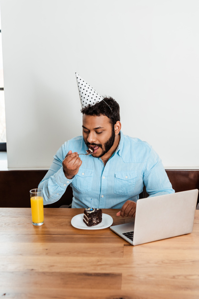 bearded african american man in party cap eating tasty birthday cake near laptop and glass of orange juice - Photo, Image