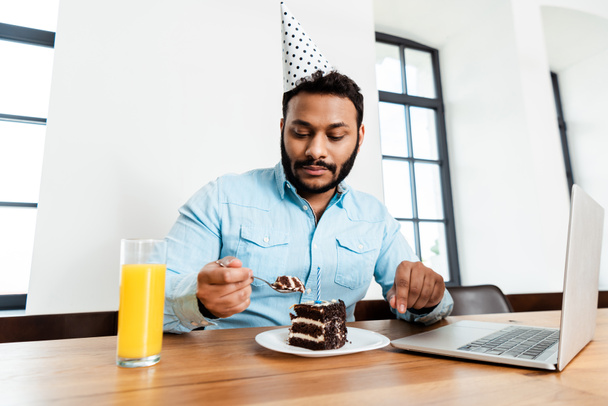 african american man in party cap looking at tasty birthday cake near laptop and glass of orange juice - Фото, зображення
