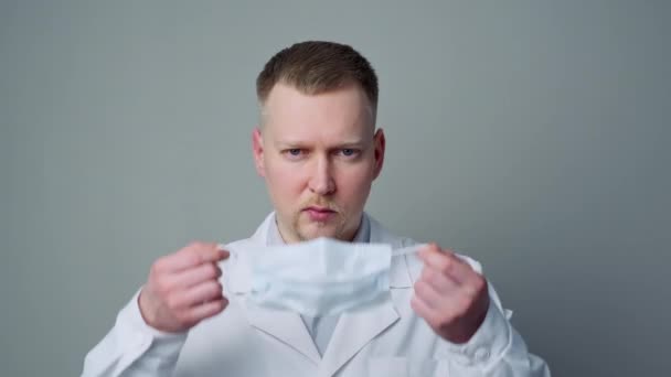 A young man doctor in a mask and a white robe on a gray background. - Materiał filmowy, wideo