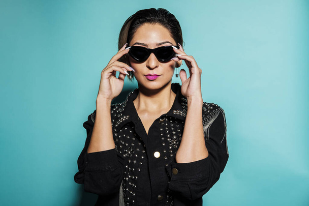 Beautiful woman portrait wearing a leather jacket and jeans with sunglasses on a blue background - Фото, зображення