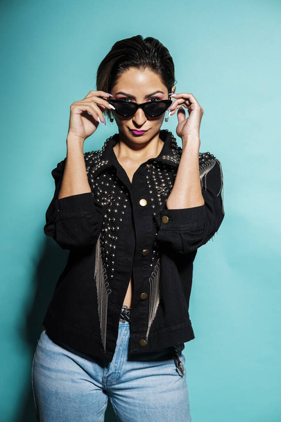 Beautiful woman portrait wearing a leather jacket and jeans with sunglasses on a blue background - Фото, изображение