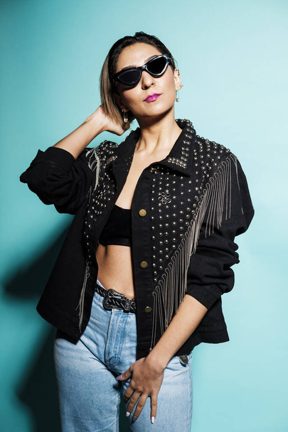 Beautiful woman portrait wearing a leather jacket and jeans with sunglasses on a blue background - Фото, зображення