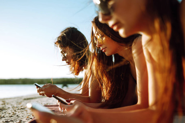 Kolme kaunista tyttöä käyttävät älypuhelinta rannalla. Tytöt ottavat aurinkoa rannalla. Nuoret tytöt tekevät selfietä puhelimen kanssa. Nauti kesäajasta
. - Valokuva, kuva