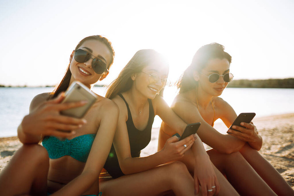 ビーチで携帯電話を使用して3人の美しい女の子。女の子はビーチで日光浴。若い女の子は電話で自撮りをしている。夏の時間を楽しむ. - 写真・画像