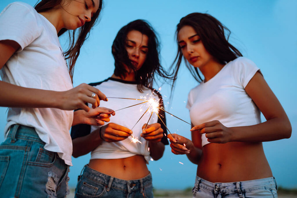 The sparklers in the hands of young girls on the beach. Three girls enjoying party on beach with sparklers. Summer holidays, vacation, relax and lifestyle concept. - Zdjęcie, obraz