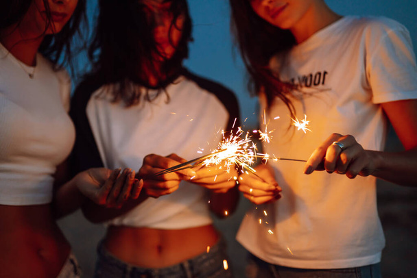 The sparklers in the hands of young girls on the beach. Three girls enjoying party on beach with sparklers. Summer holidays, vacation, relax and lifestyle concept. - Foto, Imagen