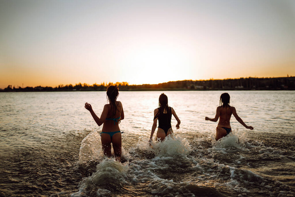 Three young girls run to swim in the sea at sunset. Summer holidays, vacation, relax and lifestyle concept. - Valokuva, kuva