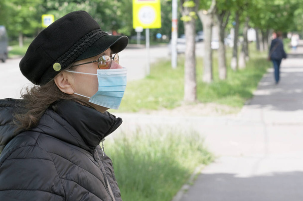Ελκυστική γυναίκα με μαύρο καπέλο με γείσο και φορώντας ιατρική μάσκα περπατώντας σε δρόμο της πόλης την ανοιξιάτικη ηλιόλουστη μέρα κοντινό πλάνο - Φωτογραφία, εικόνα