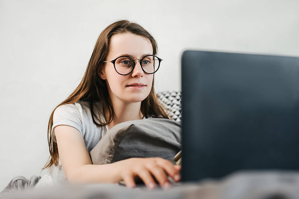 Smiling young woman in eyeglasses sitting in bed spend free time at home surfing internet. Self-employed female working using pc texting business message answering to client distantly. Selective focus - Foto, Bild