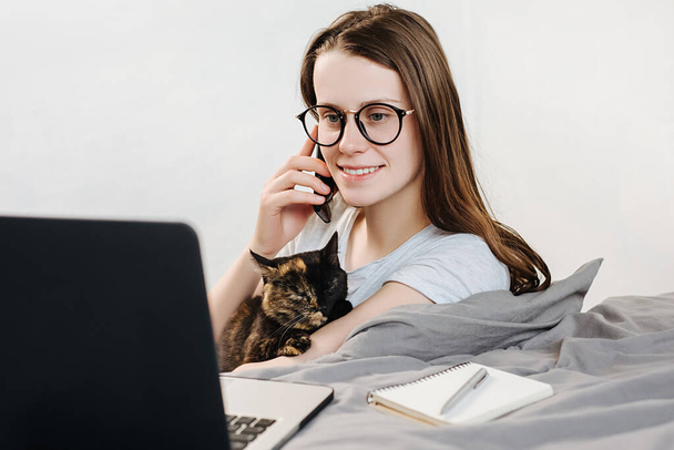 Attractive happy young woman in eyeglasses holding a cat talking by phone, making call, looking at laptop screen lying in cozy bed. Cute girl student freelancer working at home - Fotoğraf, Görsel
