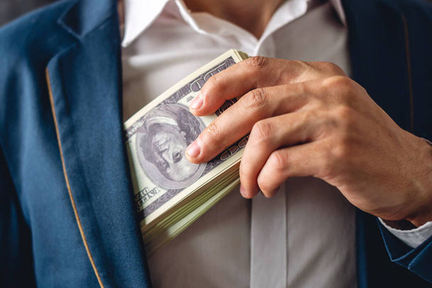 Man in a suit putting a wad of hundred-dollar bills in the inside pocket of his jacket. Close up. The concept of corruption and bribery officials - Photo, Image