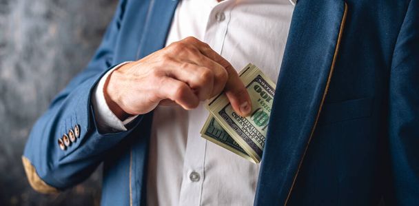 Homem em um terno colocando um wad de contas de cem dólares no bolso interior de sua jaqueta. Feche. O conceito de corrupção e funcionários do suborno - Foto, Imagem