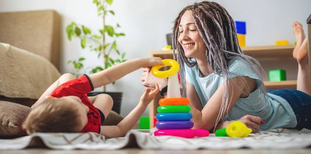 Gelukkige jonge moeder en haar kind verzamelen samen een kleurrijke piramide. Concept van spelen met kinderen, genieten van het doorbrengen van tijd thuis met het gezin. - Foto, afbeelding