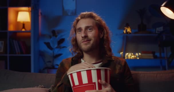 Handsome young man eating popcorn and watching film at home. Mullennial guy wit beard and long hair sitting on sofa in front of tv and smiling while spending time in evening. - Video