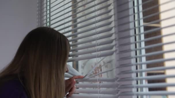 Κορίτσι με κιάλια κοιτάζει μέσα από τα στόρια από το παράθυρό του, γυναίκα κατάσκοπος - Πλάνα, βίντεο