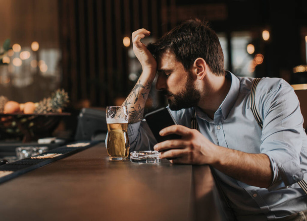 Молодой человек в беде пьет пиво, курит сигарету и пользуется мобильным телефоном в пабе
 - Фото, изображение