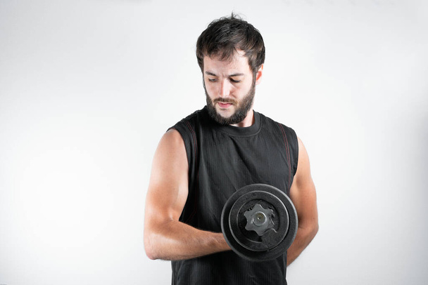 Young and handsome man with a beard in fitness session, lifting weights or dumbbells, exercising the biceps muscle. On a white background. - Foto, imagen