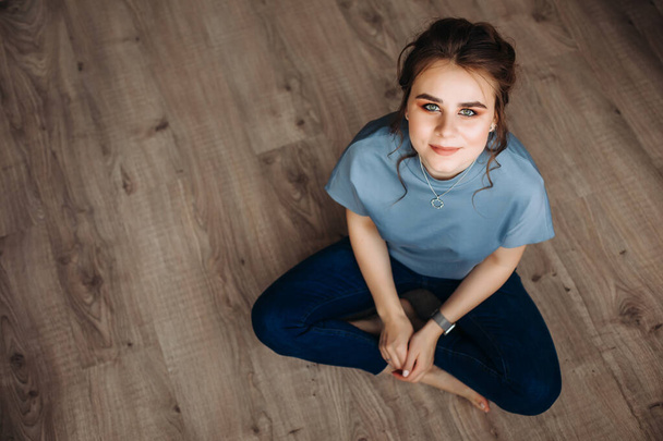 A young girl is sitting on a wooden floor, barefoot, looking at the camera. In a blue blouse and jeans. Beautiful makeup, collected hairstyle. view from above. Copy spase - Foto, Bild