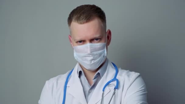 A young man doctor in a mask and a white robe on a gray background. - Záběry, video